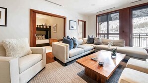 A modern living room with a beige sofa, a wooden coffee table, and a large window with a view of a snowy landscape. - Solaris Residences 5C West (Vail)