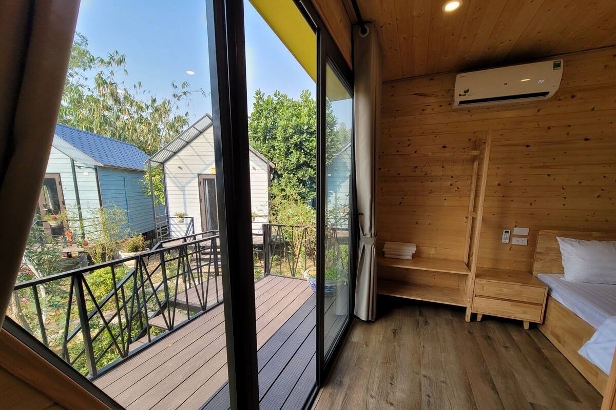 A room with a wooden floor, a bed with white bedding, a small bedside table, a wall-mounted air conditioner, and a view of a deck and outdoor area with plants and a blue-roofed structure.