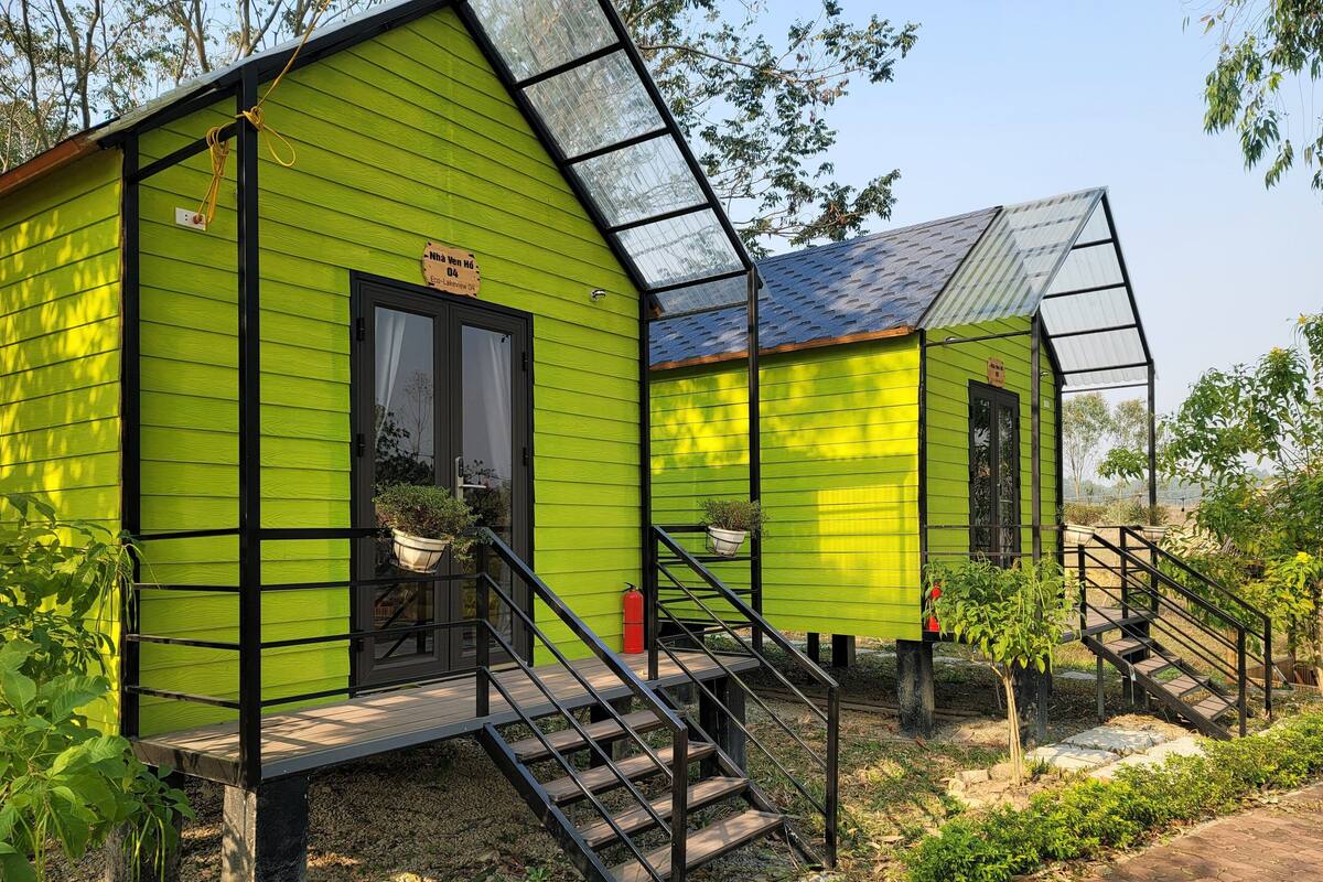 A green cabin with a staircase leading to a door, surrounded by greenery.