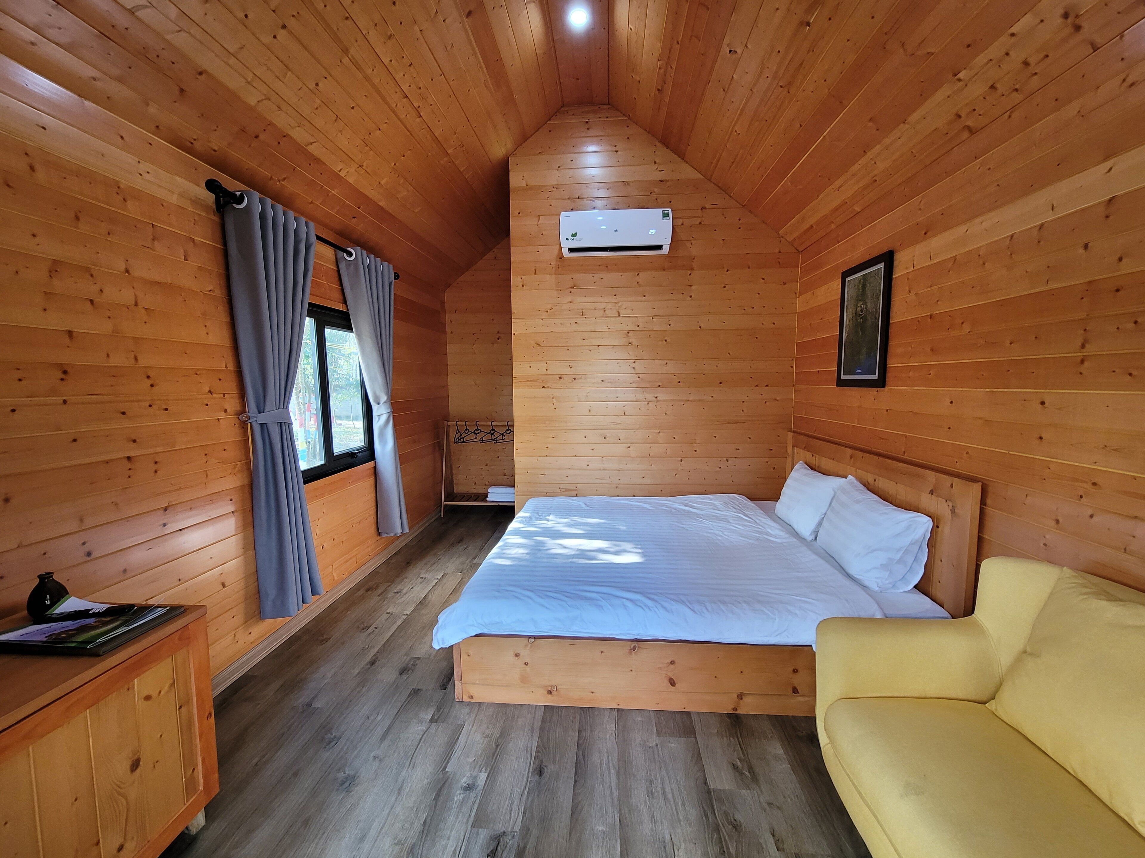 Une pièce de cabane en bois comprenant un lit, un canapé jaune, une armoire en bois et une fenêtre avec des rideaux.