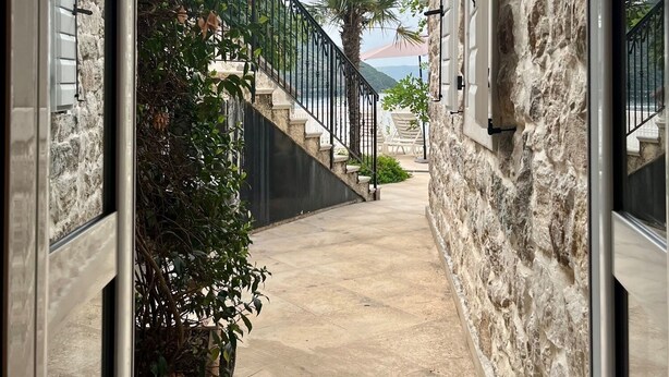 A narrow outdoor corridor with stone walls, a palm tree, and a view of the sea.