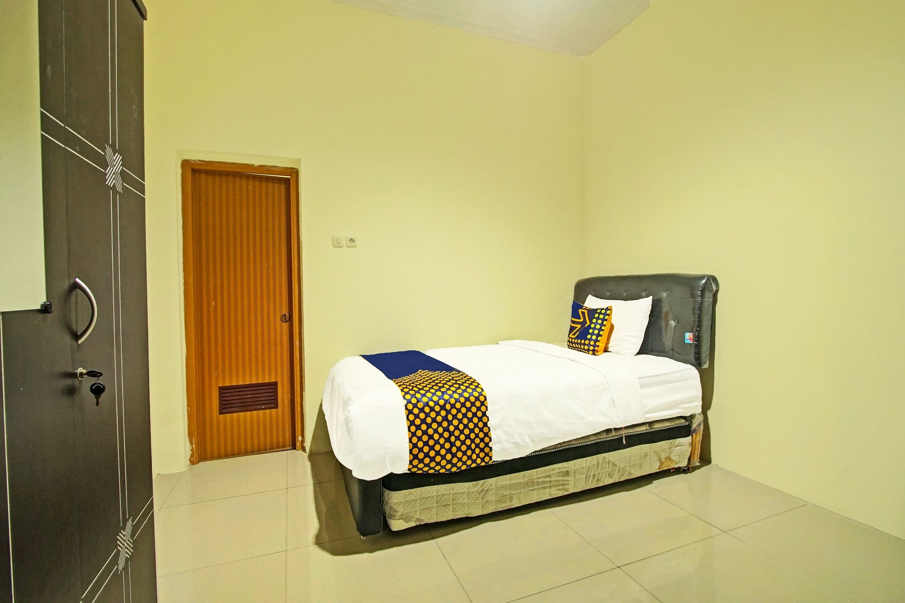A single bed with a white mattress, a patterned blanket, and a pillow with a geometric design. A wooden door with a window is visible to the left.