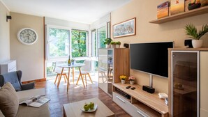 Un salon moderne doté d’un téléviseur à écran plat, de meubles en bois, d’un coin repas avec une table et des chaises, et d’une grande fenêtre offrant une vue sur la verdure.