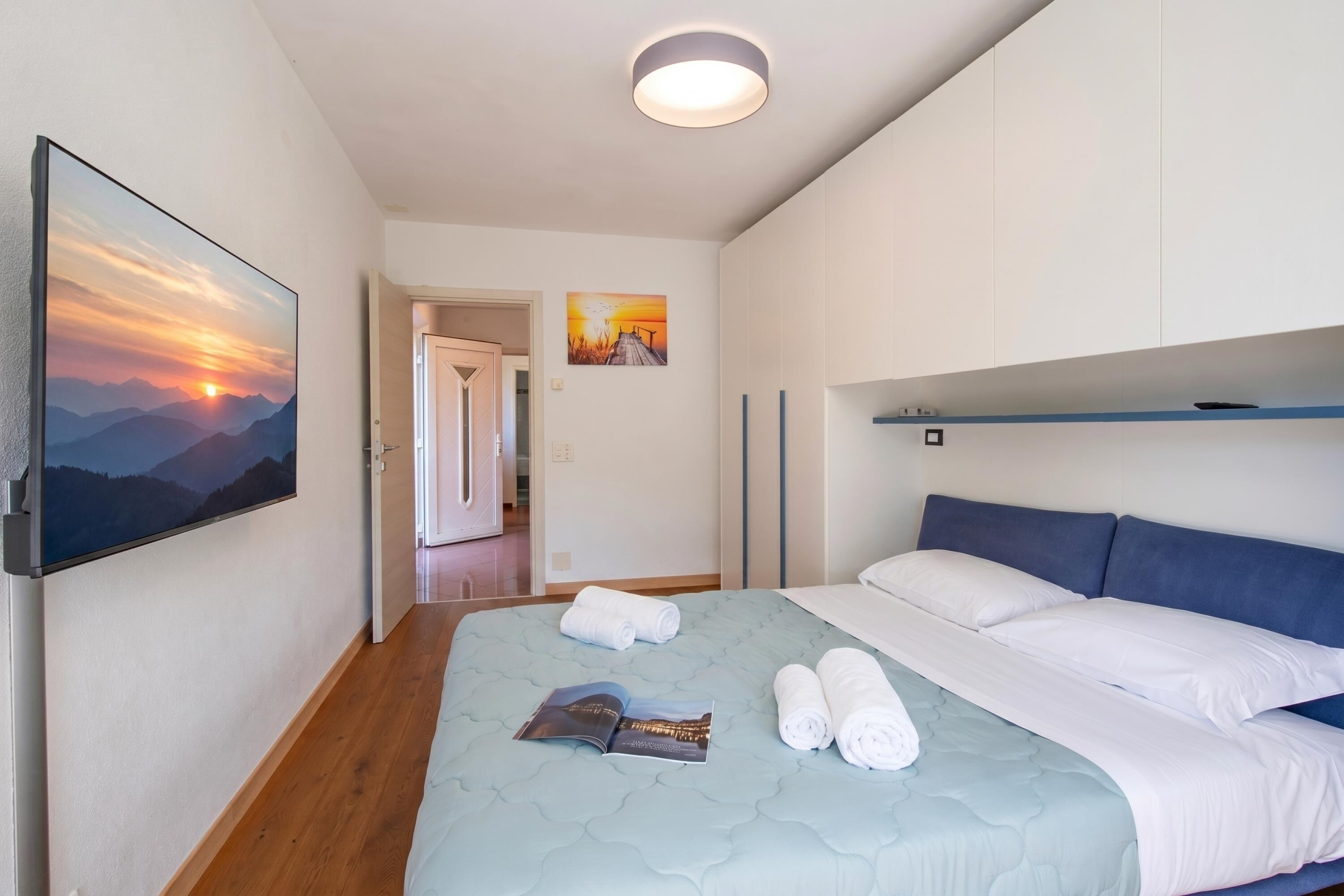 Habitación de hotel con cama, un cuadro de un atardecer, una puerta que da a otra habitación y una mesita con un libro y toallas.
