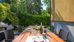 Une table en bois installée à l’extérieur, prête pour un repas, avec des chaises, une bouteille de vin et une tasse.