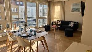 A modern living room with a dining area, large windows, a black sofa, and a small table with a vase. - Seashore (Koksijde)