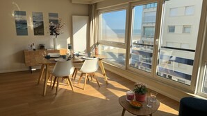 A modern dining area with a wooden table and chairs, a small side table with a vase and glass, and large windows offering a view of the cityscape. - Seashore (Koksijde)