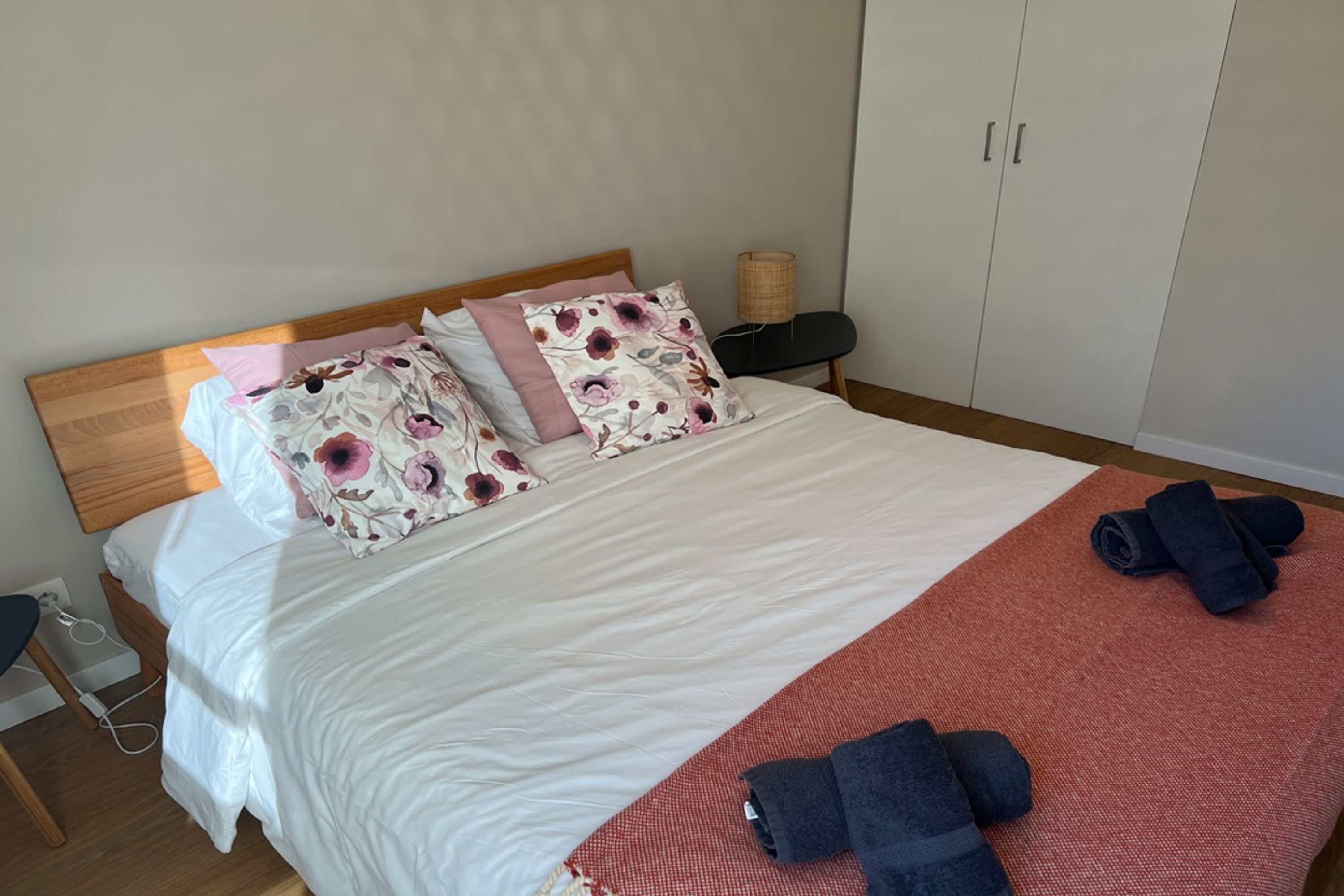 Ein ordentlich bezogenes Bett mit blumigem Kissenmuster, einer roten Tagesdecke und aufgerollten Handtüchern auf Holzdielen.