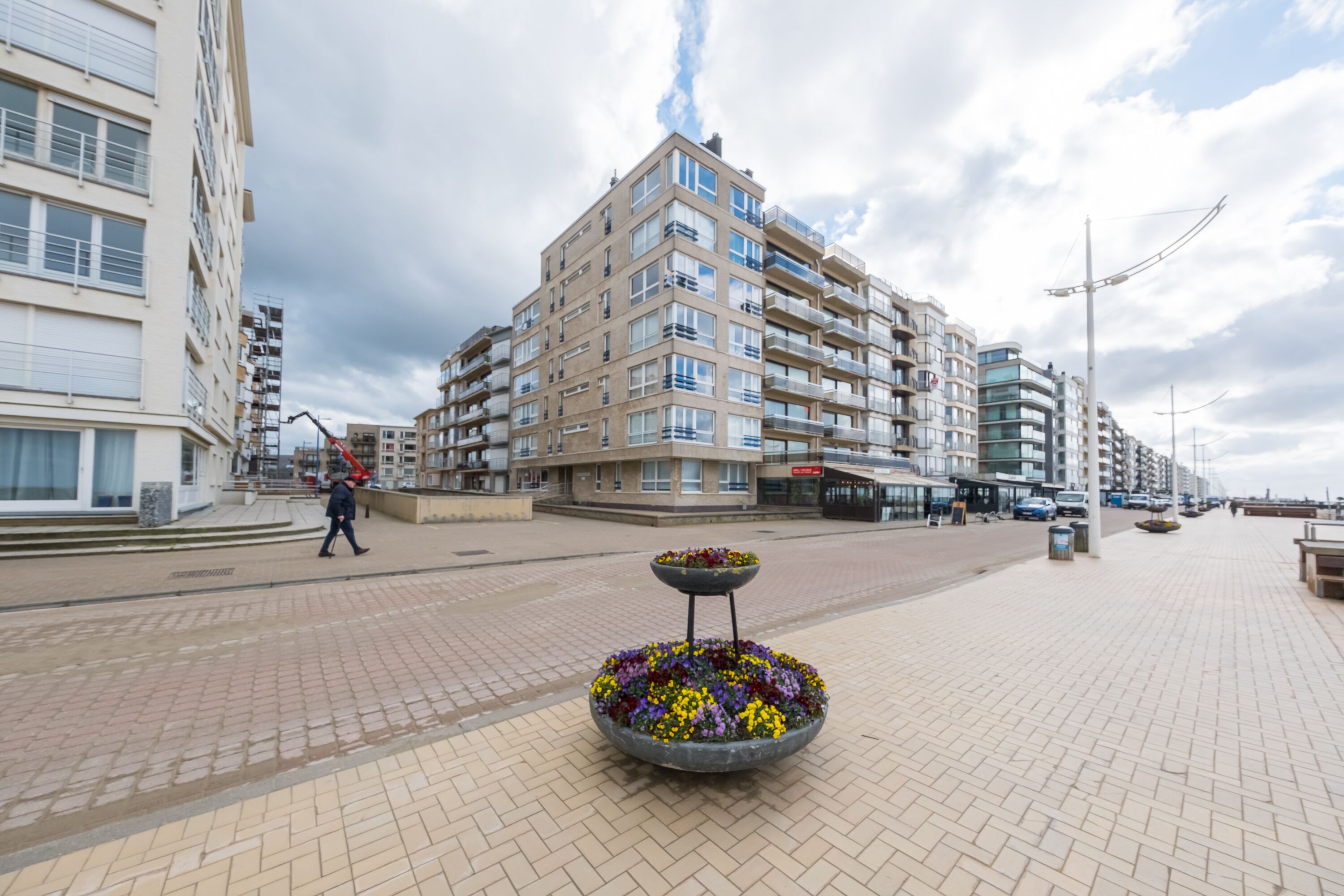Een meerlaags gebouw met balkons, een geplaveid voetpad en een bloembed in de voorgrond.