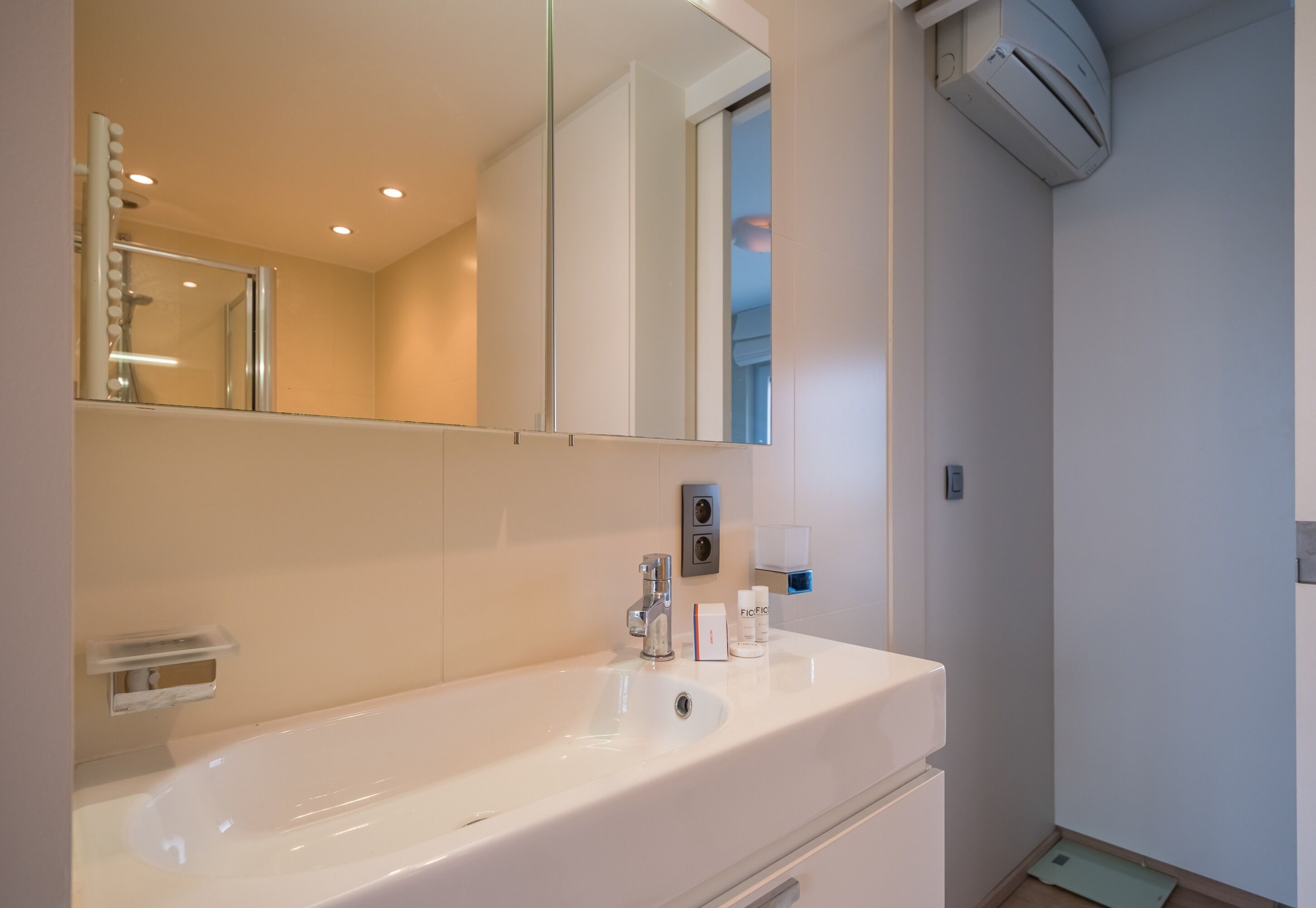 A bathroom with a white sink, mirror, and shower. There are toiletries on the sink and an air conditioning unit on the wall.