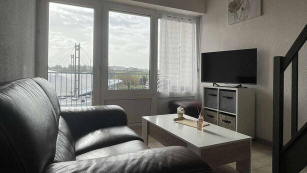 A living room with a black leather sofa, a white coffee table, a flat-screen TV on a cabinet, and a large window with a view of a bridge and buildings. - 3-room apartment for 4 in Châtelaillon-Plage with parking (Châtelaillon-Plage)