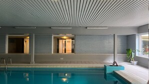 A swimming pool with a tiled deck, a ceiling with a linear pattern, and a window with a view of the outside. - Westend Palace (Middelkerke)