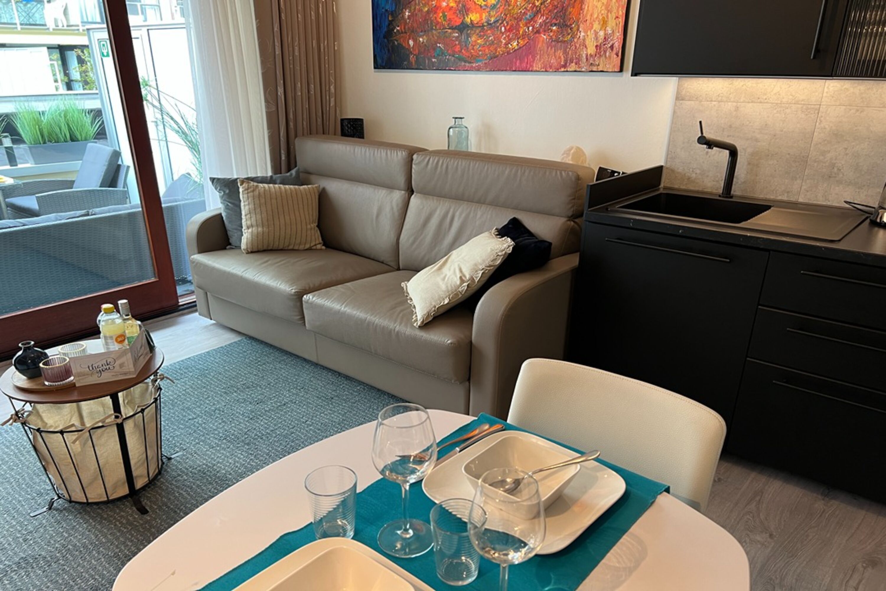 A modern hotel room with a beige sofa, a dining table with a teal placemat, and a kitchen area with a black sink and cabinets.
