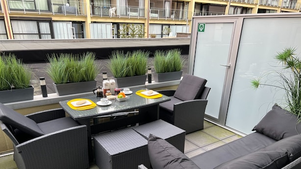 An outdoor seating area with wicker furniture, a glass-top table, and potted plants. - Westend Palace (Middelkerke)