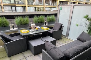 Un área de estar al aire libre con mobiliario de mimbre, una mesa con superficie de vidrio y plantas en macetas.