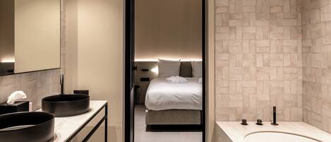 A modern bathroom with a large bathtub, two round sinks, and a bed visible through an open doorway.