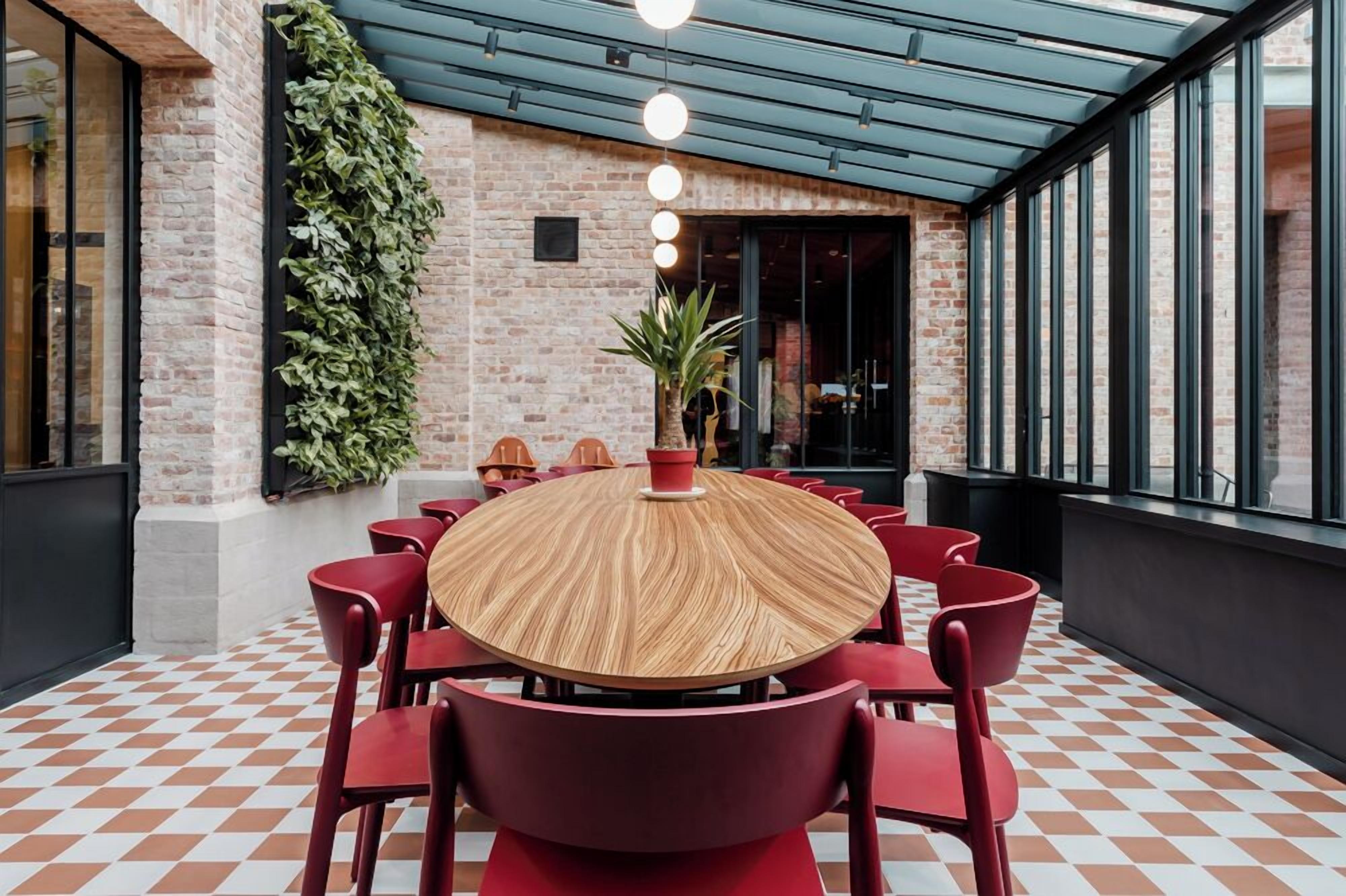 Una mesa redonda de madera rodeada de sillas rojas en una habitación con piso a cuadros, paredes de ladrillo y un ventanal grande.