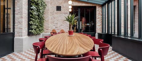 Una mesa redonda de madera rodeada de sillas rojas en una habitación con piso a cuadros, paredes de ladrillo y un ventanal grande.