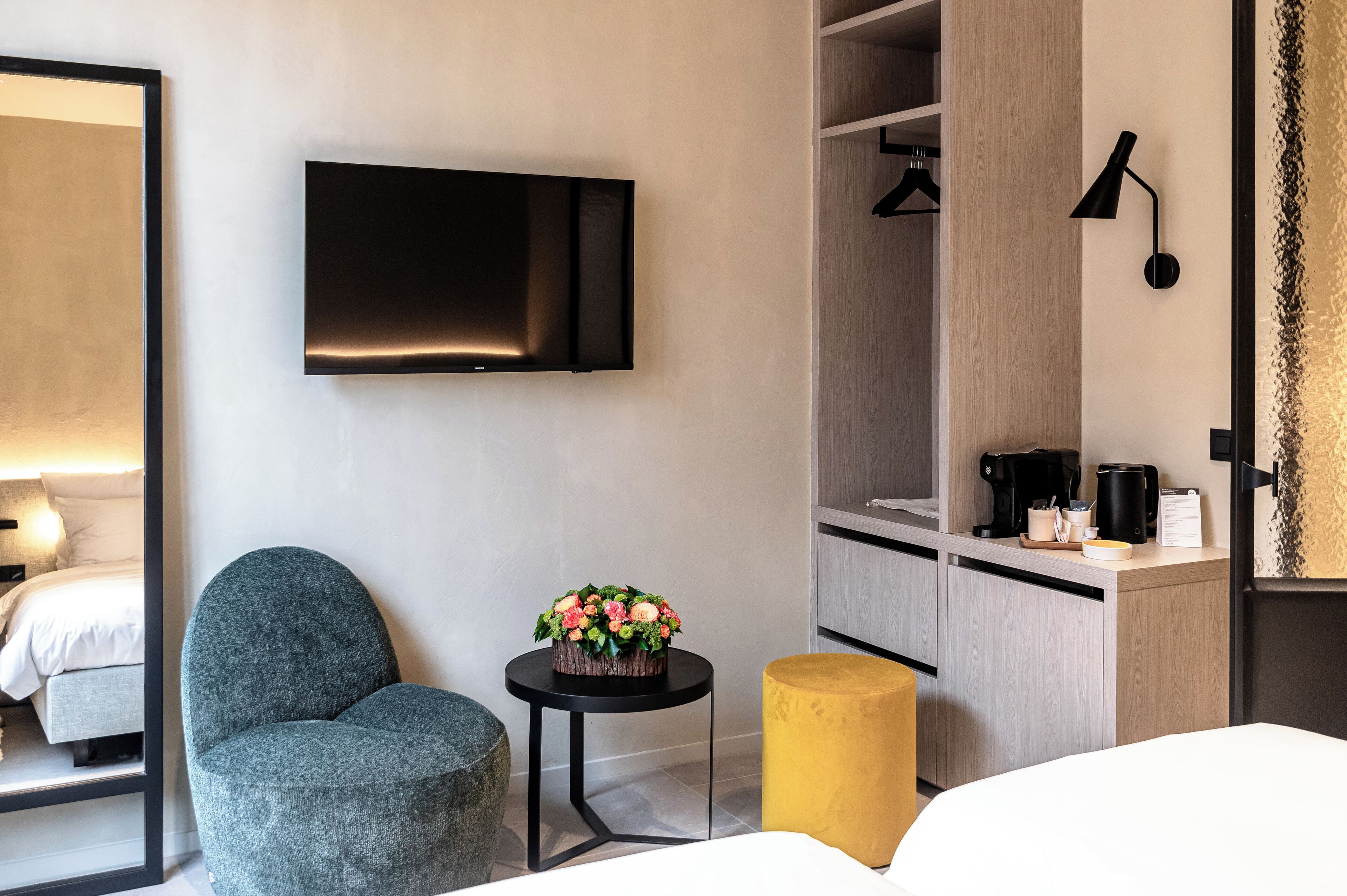 A modern hotel room with a bed, a grey armchair, a small table with a flower arrangement, a yellow ottoman, a TV mounted on the wall, a wardrobe, and a coffee machine.