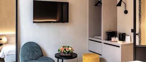 Une chambre d’hôtel moderne comprenant un lit, un fauteuil gris, une petite table agrémentée d’un arrangement floral, un pouf jaune, un téléviseur fixé au mur, une armoire et une machine à café.