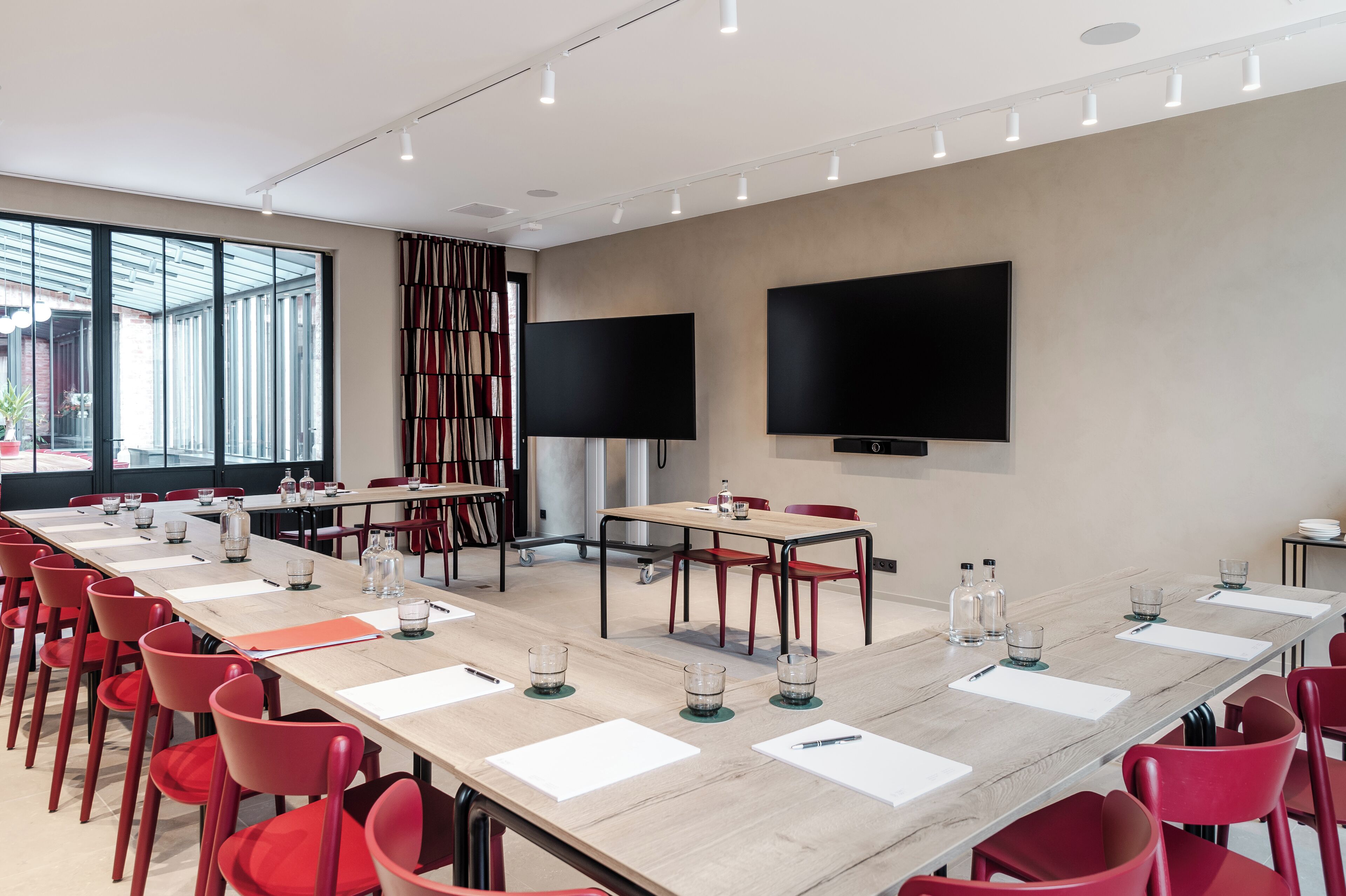 A conference room with a long table, red chairs, and two large screens on the walls.