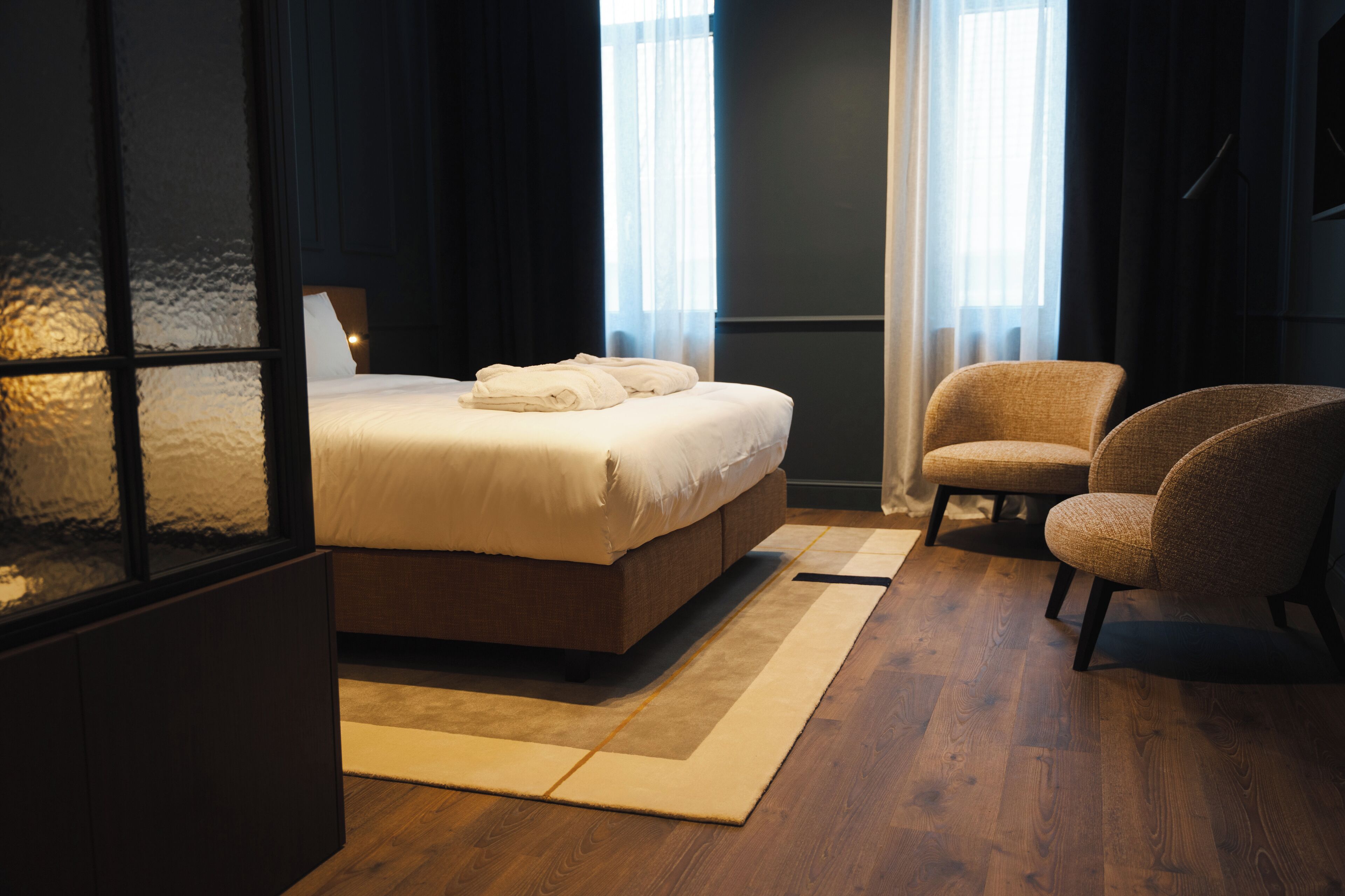 A modern hotel room with a large bed, a chair, and a view of the city through the window.