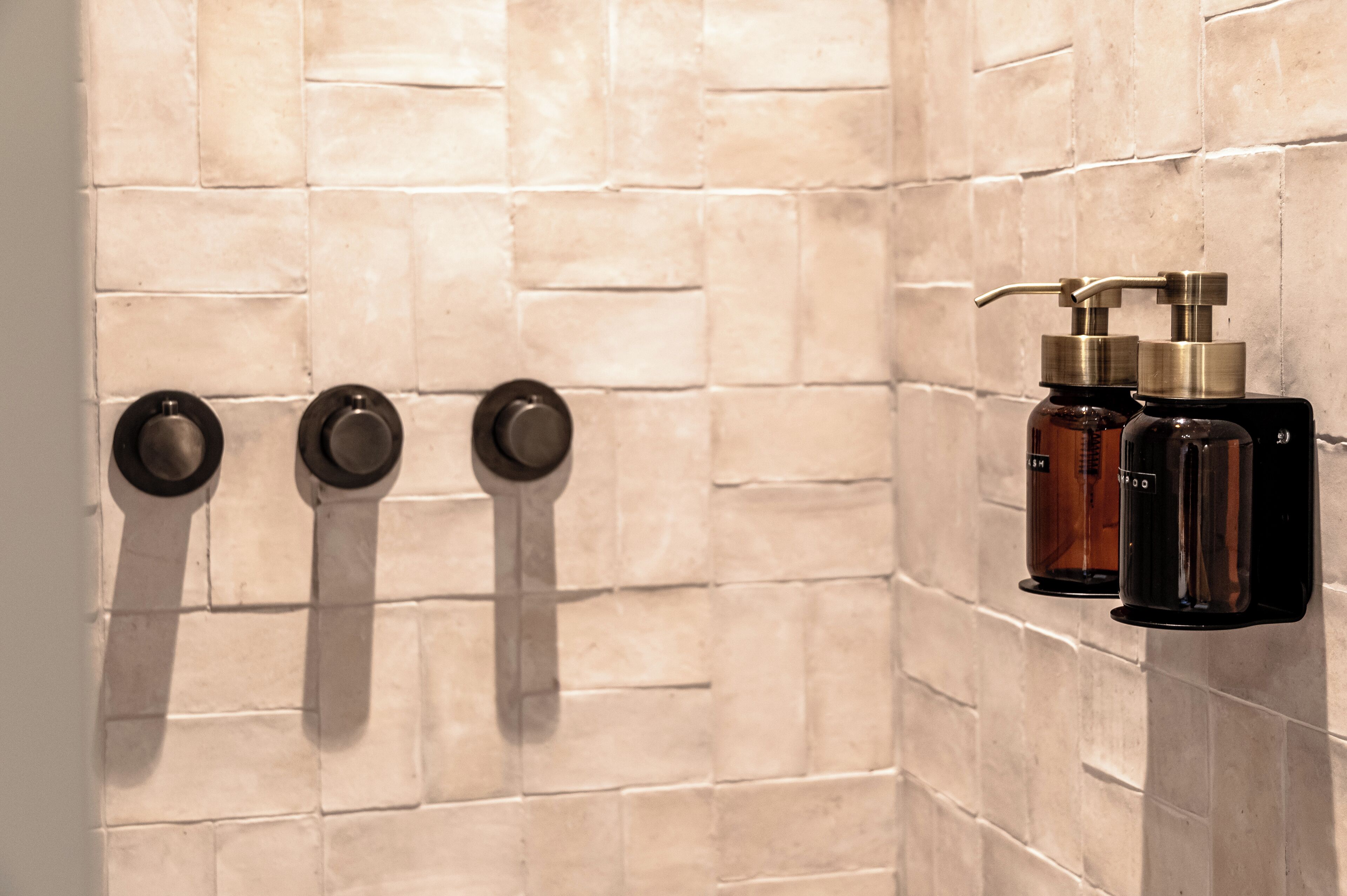 Un baño con paredes de azulejos color beige y un perchero negro con tres toallas. Hay dos botellas de artículos de aseo en un estante negro.