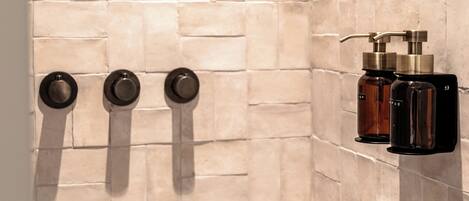 Un baño con paredes de azulejos color beige y un perchero negro con tres toallas. Hay dos botellas de artículos de aseo en un estante negro.