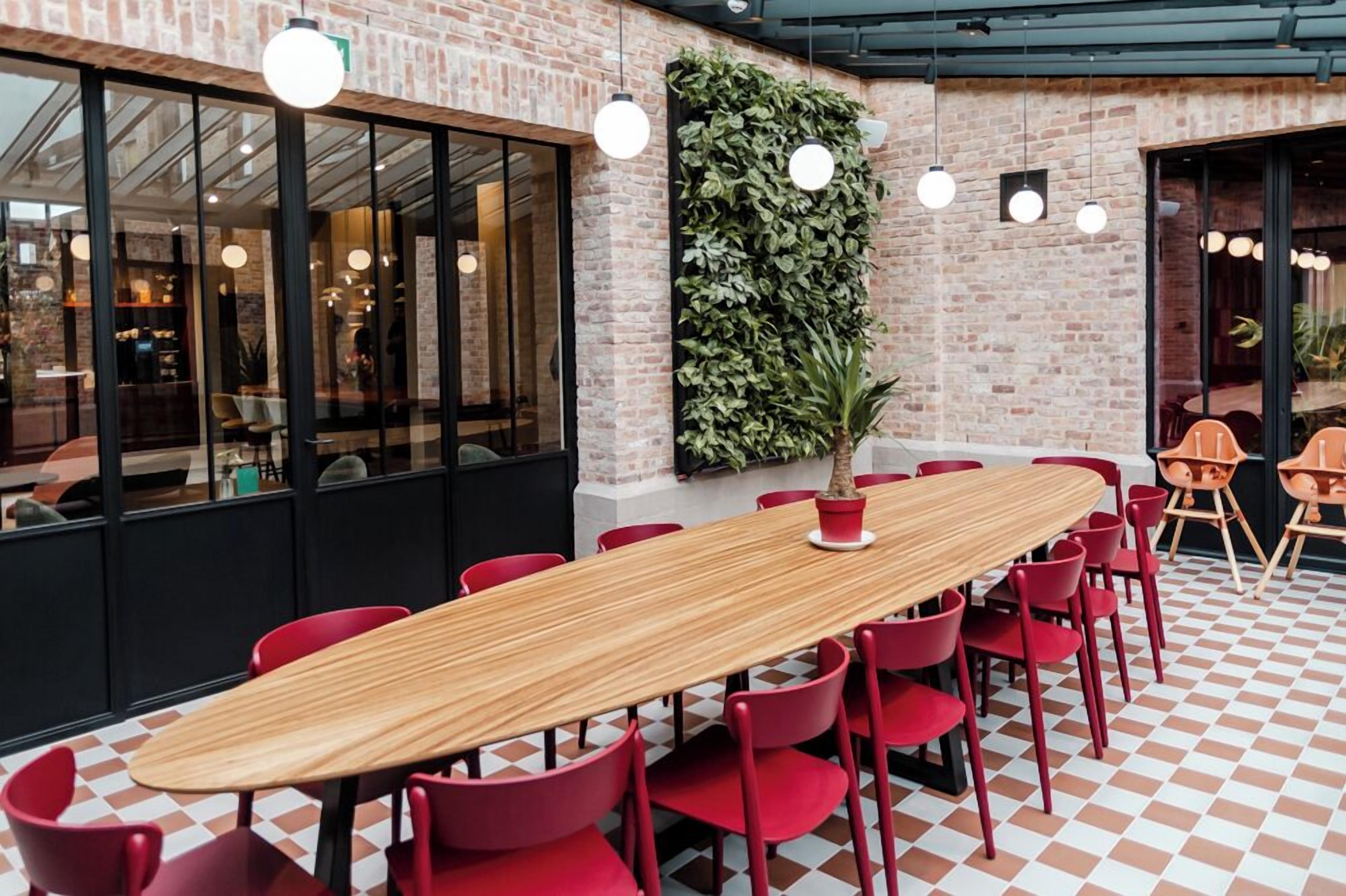 A long wooden table with red chairs in a modern indoor space with brick walls and large windows.