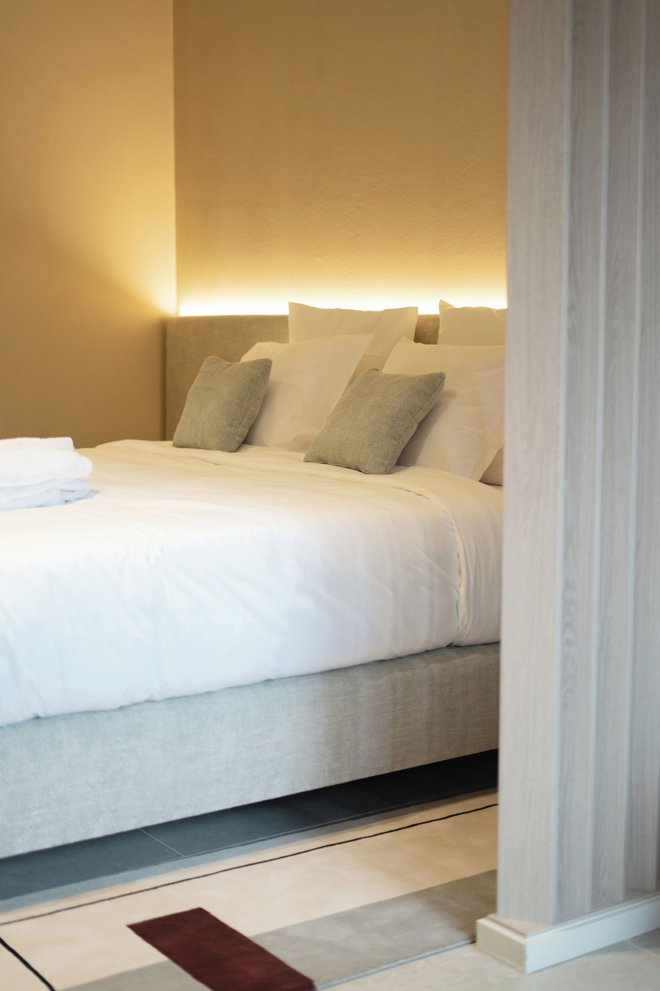Una cama bien hecha con sábanas blancas y una alfombra marrón decorativa, un cabecero y una lámpara fijada a la pared.