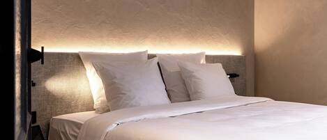A neatly made bed with white linens and pillows, a headboard with built-in lighting, and a dark-colored chair beside the bed.