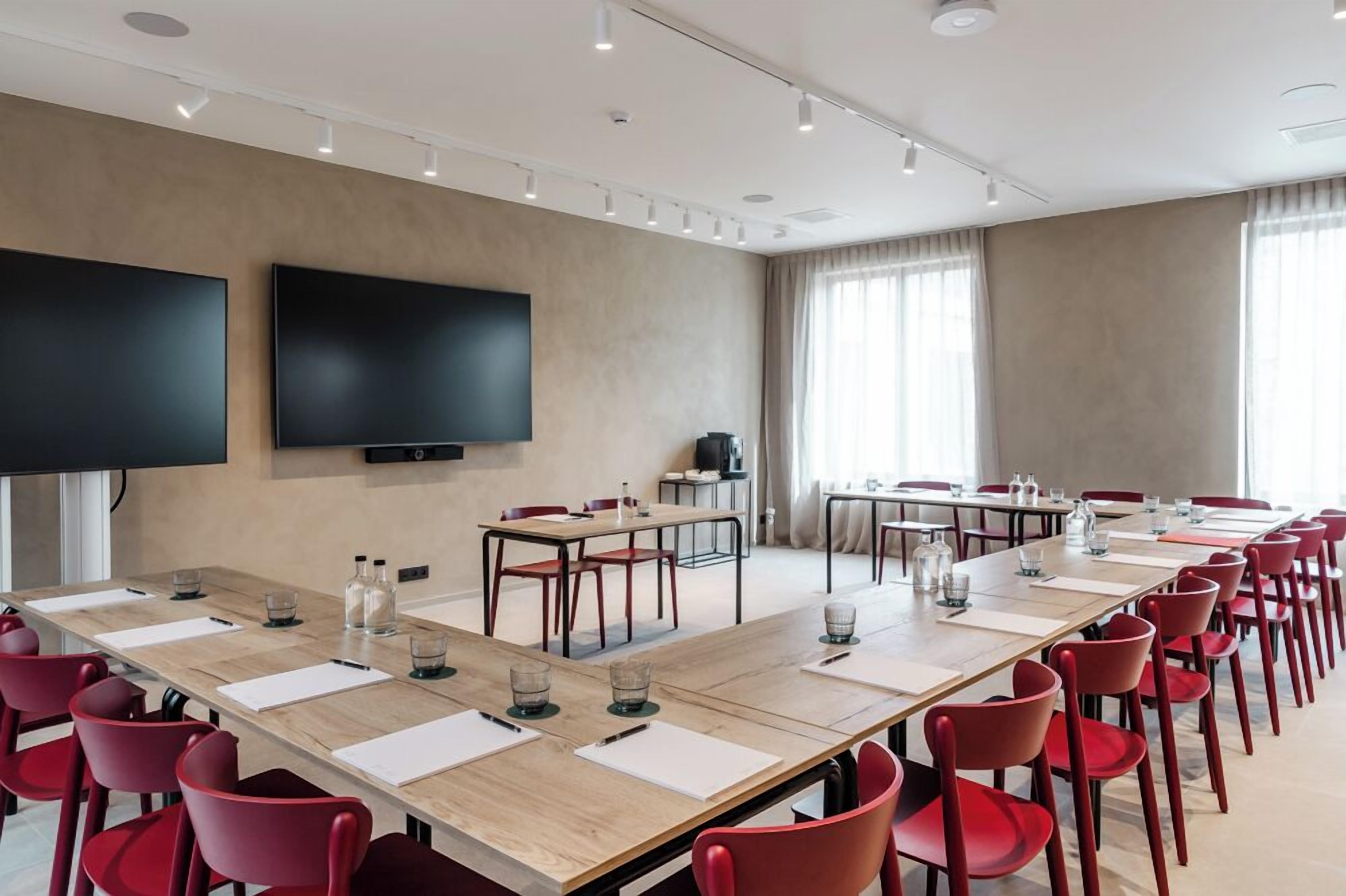 Ett konferensrum med ett långt bord, röda stolar, en platt-TV och ett stort fönster med gardiner.