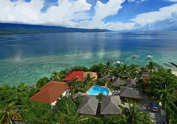 Un resort con piscina rodeada de cabañas con techo de paja, palmeras y vistas al océano.