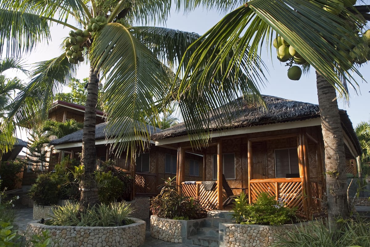 Un resort tropical con una edificación de techo de paja, macetas de piedra y exuberante vegetación.