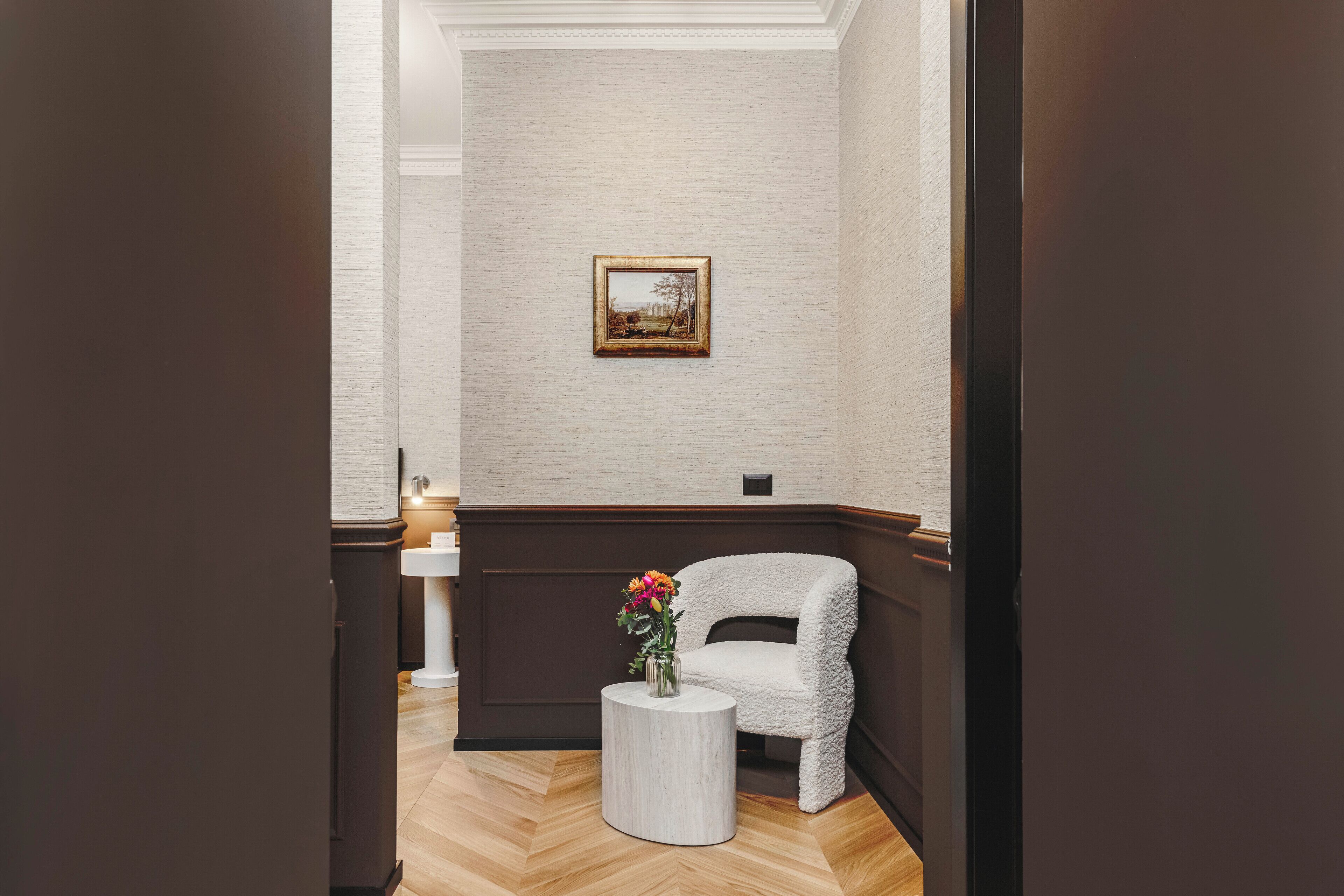 A hallway with a white chair, a small round table with a vase of flowers, and a framed picture on the wall.