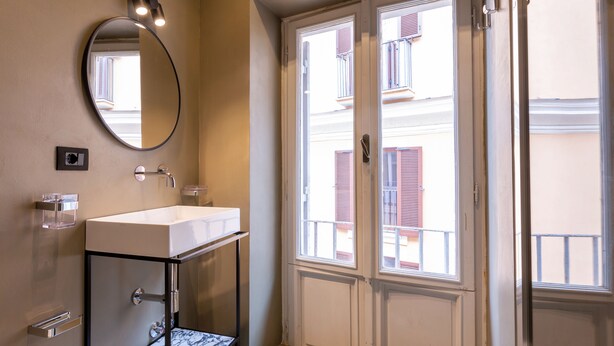 A bathroom with a white sink, a mirror, and a window with a view of buildings. - The Couper Colonna (Rome)