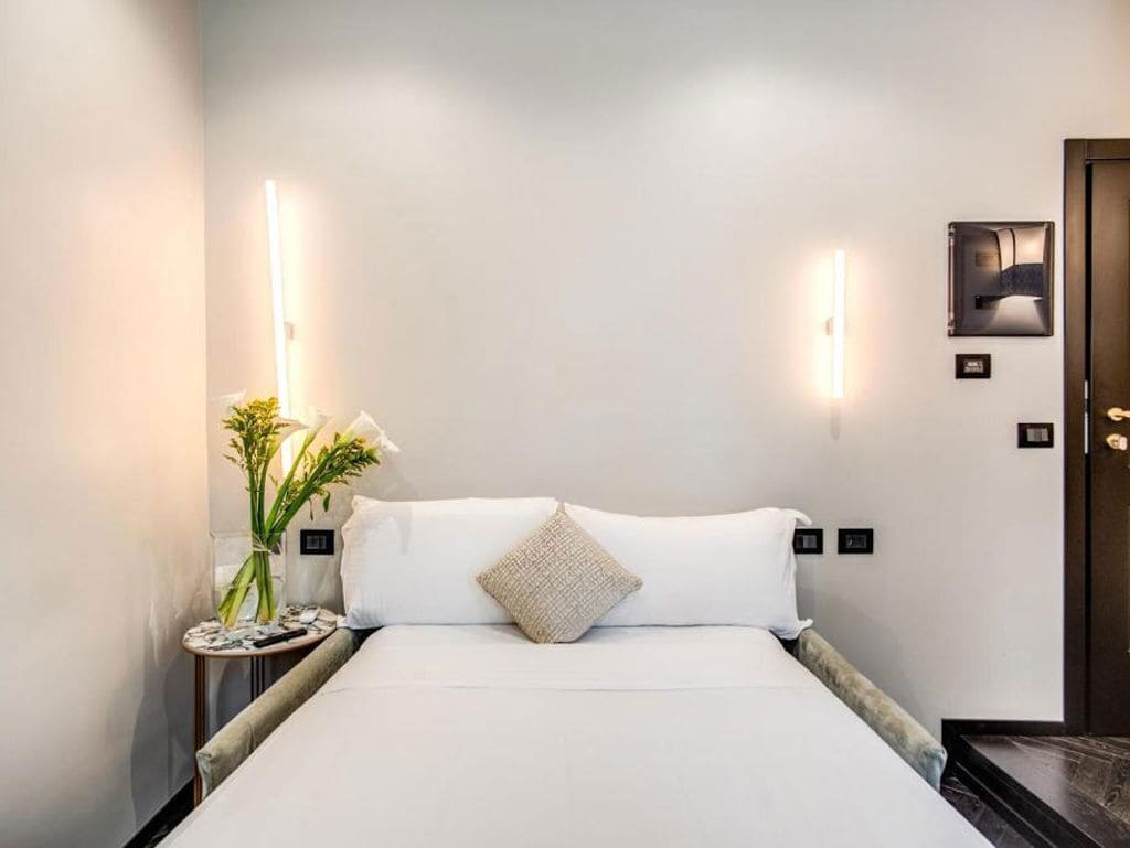 A neatly made bed with white linens and a single decorative pillow, flanked by two wall-mounted light fixtures.