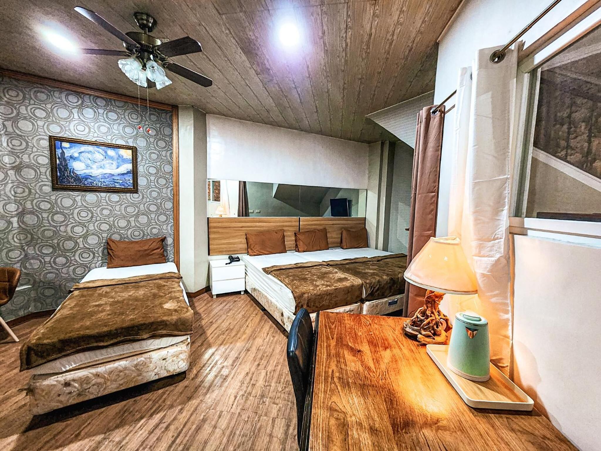 Une chambre d’hôtel avec deux lits, un plafond en bois, un ventilateur de plafond, un tableau, une table de chevet, une lampe et une fenêtre avec des rideaux.