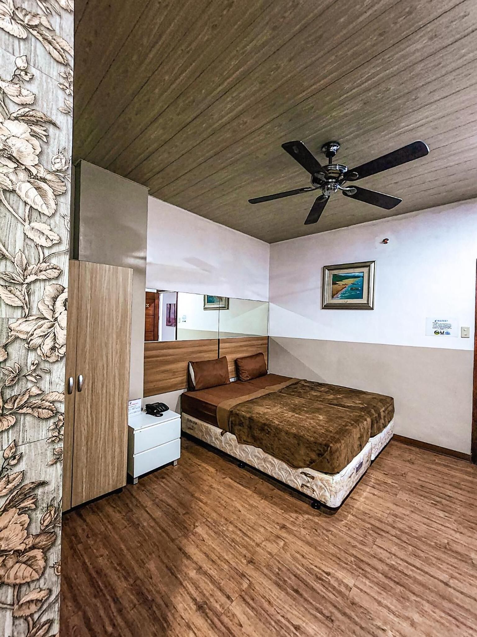 Ein Schlafzimmer mit Holzboden, einem Bett mit brauner Decke, einem Nachttisch, einem Kleiderschrank und einem Deckenventilator.