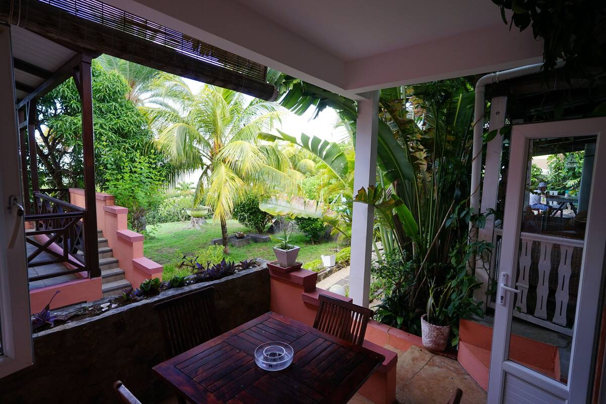 A covered outdoor area with a wooden table and chairs, surrounded by lush greenery and a view of a garden with palm trees.