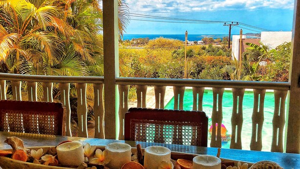 A wooden boat filled with candles and flowers on a balcony with a view of the ocean and palm trees. - Bellevue Tamarin (Tamarin)