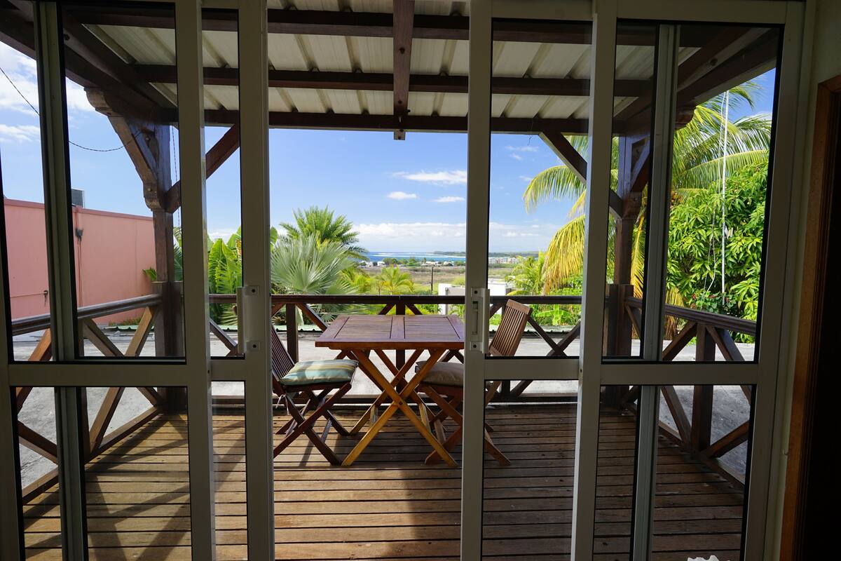 A wooden deck with a table and chairs, overlooking a scenic view with palm trees and a distant landscape.