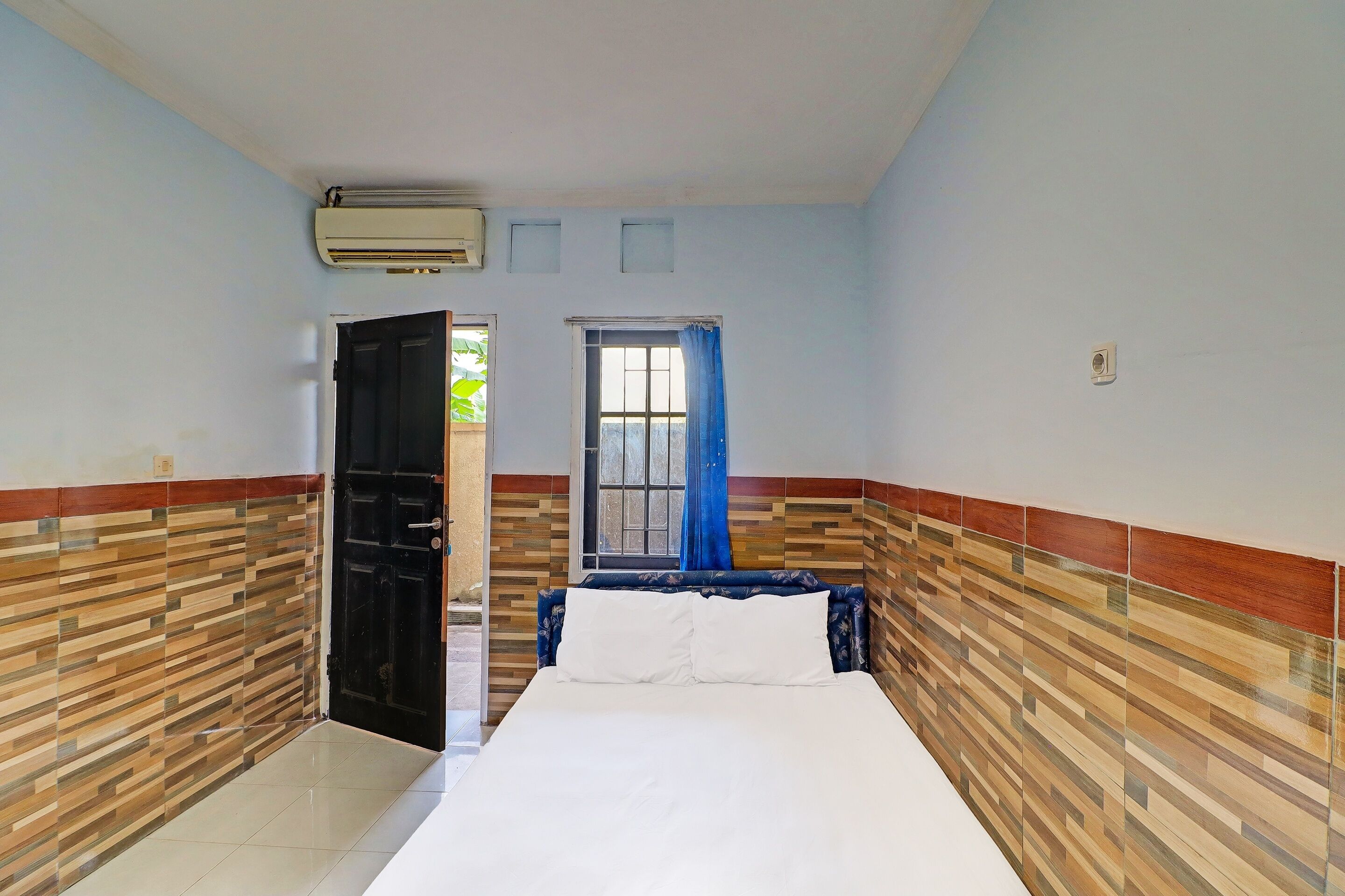 A bedroom with a bed, a window with blue curtains, a door, and a wall with a patterned design.