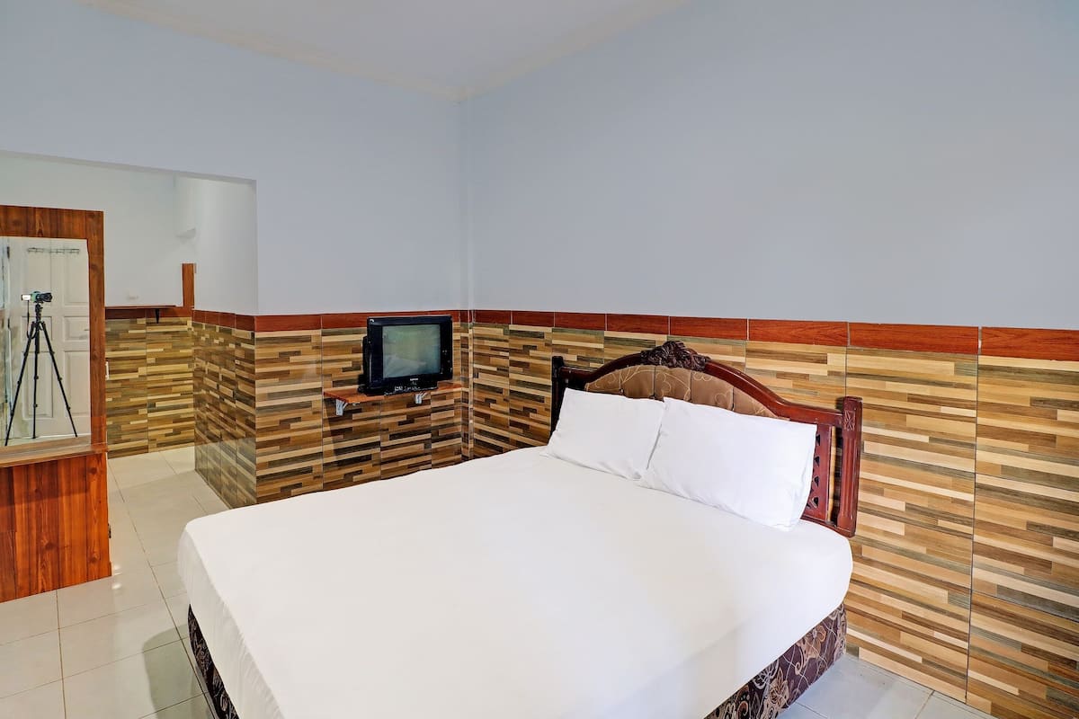 Habitación de hotel con una cama grande, cabecera de madera, televisor sobre un soporte y una cámara sobre un trípode.