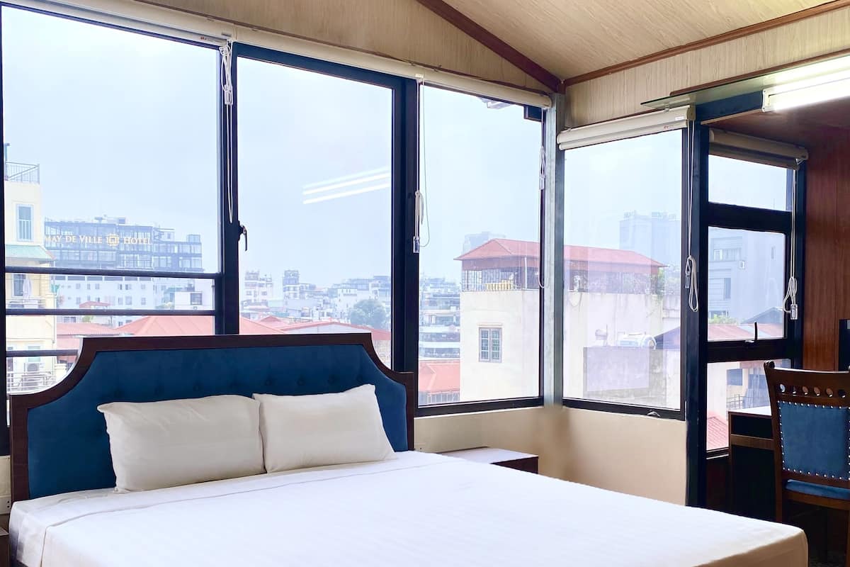 A hotel room with a large bed, a wooden headboard, and a view of the city through large windows.