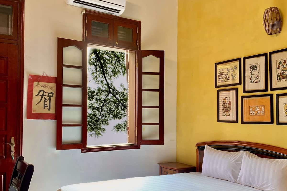 A neatly made bed in a room with a wooden headboard, a window with wooden shutters, and several framed artworks on the wall.