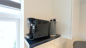 Une machine à café est posée sur un plan de travail noir, à côté d’un mur blanc, et une fenêtre se trouve à gauche.