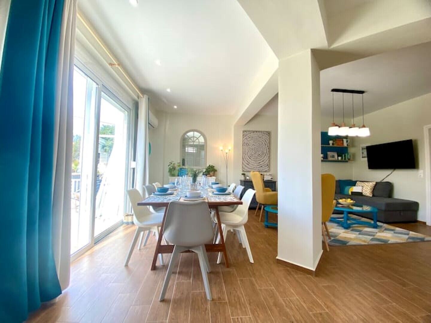 A spacious living area with a dining table set for four, a sofa, a TV mounted on the wall, and a large window with curtains.