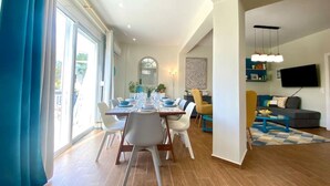 A spacious living area with a dining table set for four, a sofa, a TV mounted on the wall, and a large window with curtains. - CostAnita Beach House in Almyrida (Almirida)