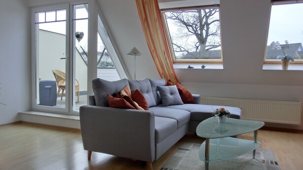 Ein modernes Wohnzimmer mit einem grauen Sofa, einem Glastisch und Blick ins Freie durch große Fenster.